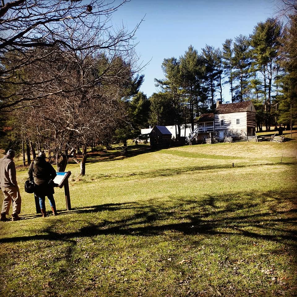 Two people stand near a Civil War Trails sign in a field surrounded by trees, a rustic small house stands in the distance